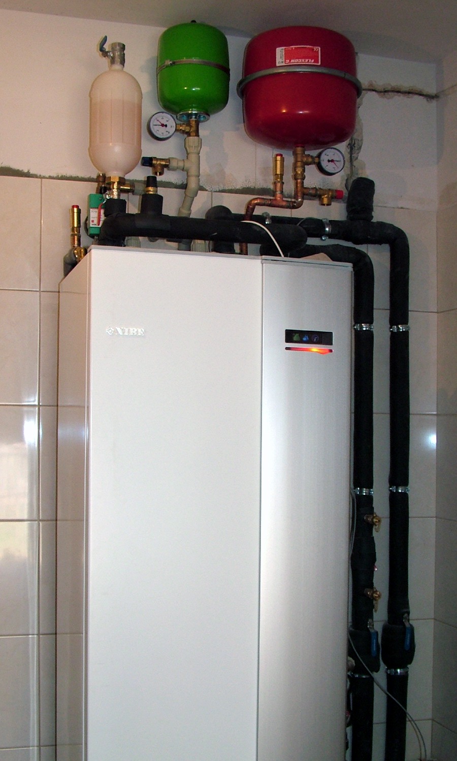 Biała pompa ciepła NIBE z zielonym i czerwonym zbiornikiem wyrównawczym, czarne rury izolowane, na tle ściany z jasnych płytek. Widoczne manometry i zawory.