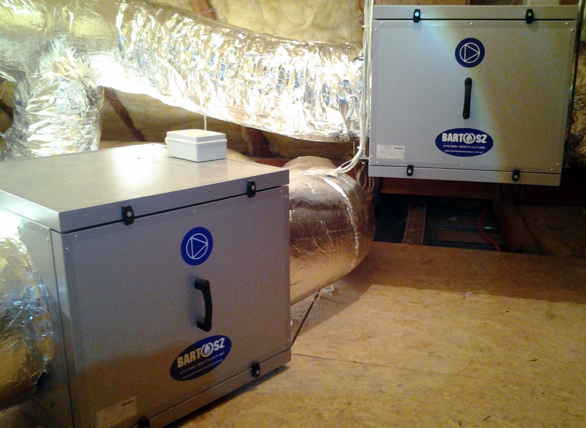 Szara jednostka rekuperacji z widocznymi przewodami wentylacyjnymi w izolacji, zamontowana w pomieszczeniu technicznym. Widoczne logo firmy.