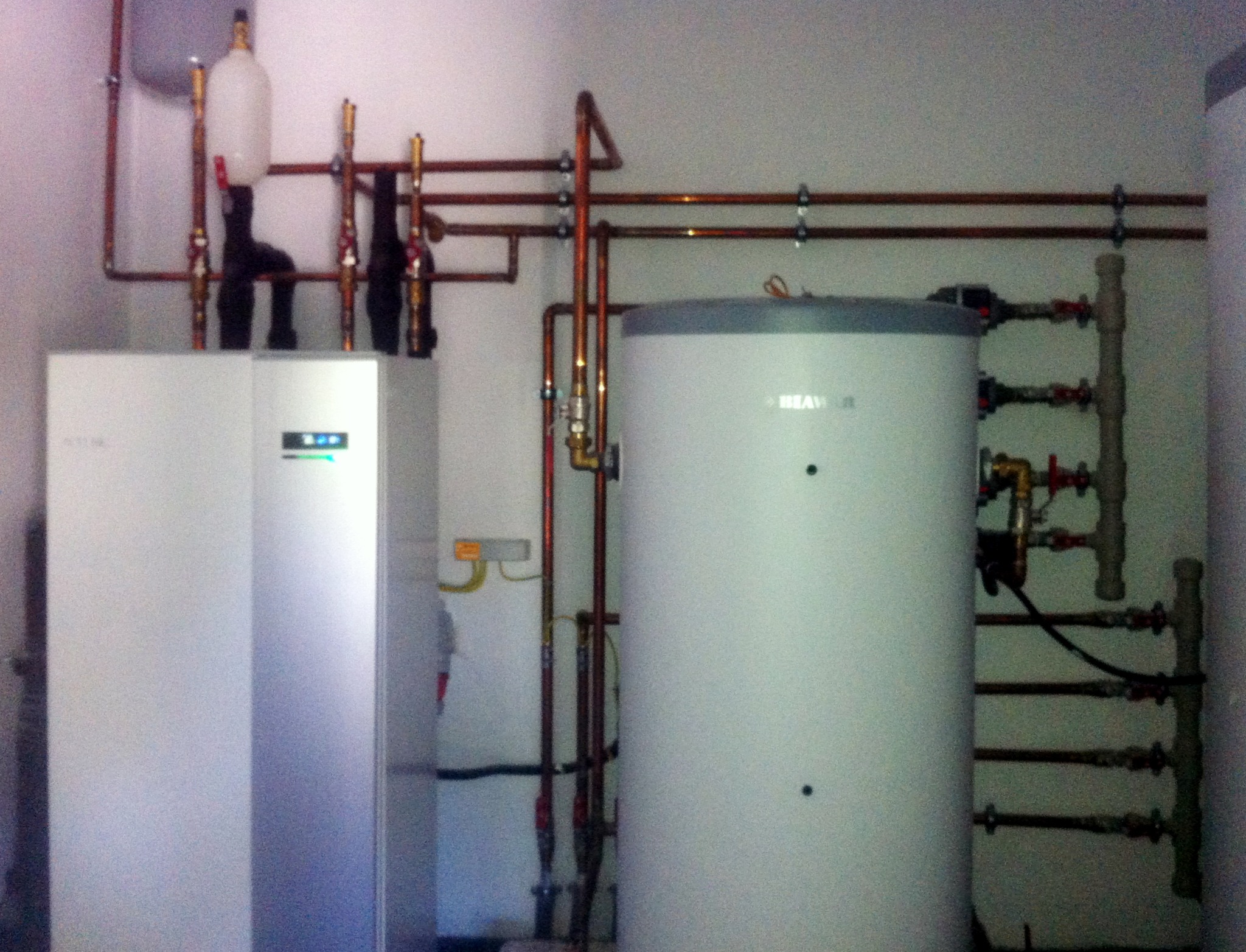 Biała pompa ciepła z zbiornikiem i miedzianą instalacją w pomieszczeniu. Widoczne liczne rury, zawory i elementy montażowe. Całość sprawia wrażenie nowej instalacji.