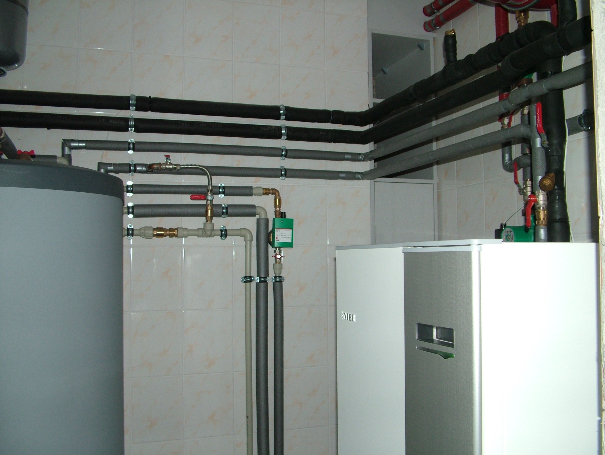 Węzeł cieplny w kotłowni: zbiornik, rury w izolacji, zawory, pompa obiegowa i nowoczesny piec NIBE na tle jasnych, kwadratowych płytek ściennych.