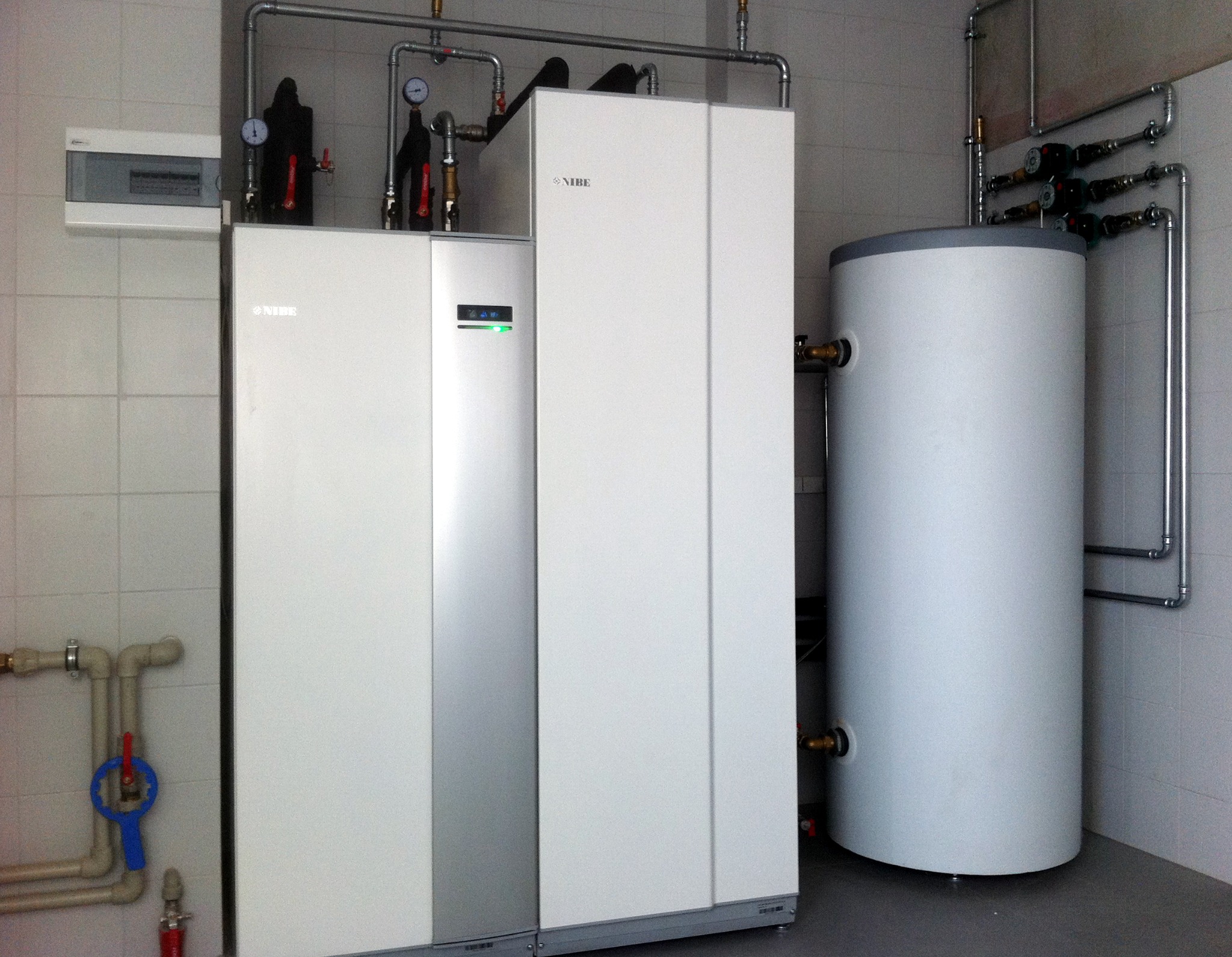 Biała, nowoczesna pompa ciepła Nibe z zbiornikiem i instalacją w pomieszczeniu z białymi płytkami. Widoczne rury i manometry.