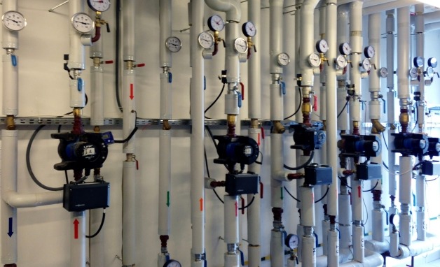 Rzędy białych rur instalacyjnych z manometrami i pompami obiegowymi, zamontowane na ścianie, z widocznymi strzałkami kierunku przepływu.
