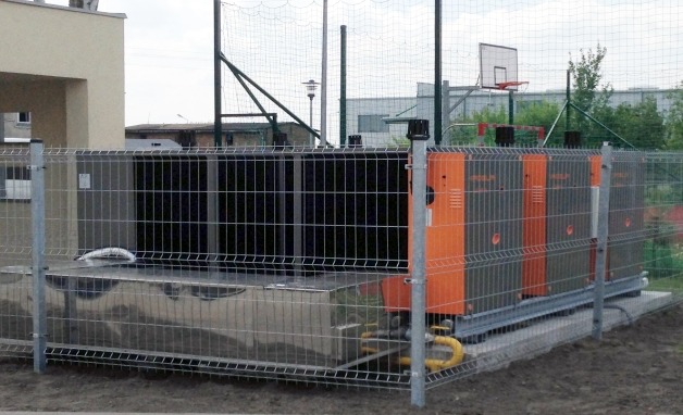 Kompleksowa instalacja przemysłowej pompy ciepła, zabezpieczona metalowym ogrodzeniem na zewnątrz budynku. Widoczne elementy instalacji i rury doprowadzające.