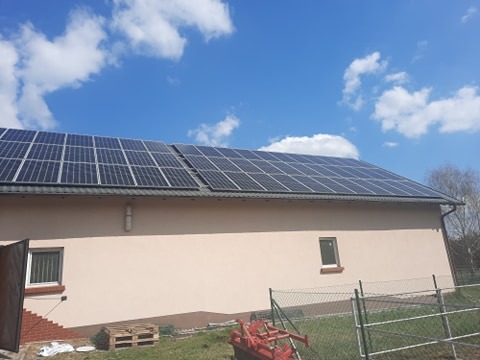 Panel słoneczny na dachu budynku o jasnej elewacji. Widoczne niebieskie niebo z białymi chmurami. W tle drzewa i ogrodzenie. Na pierwszym planie sprzęt rolniczy.