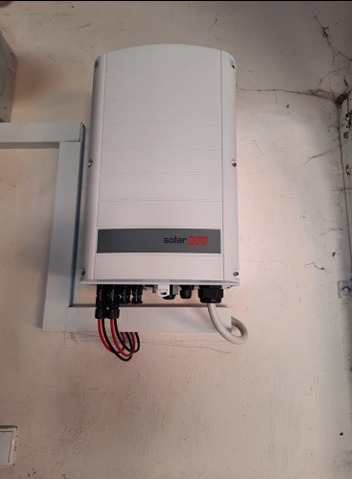 Biała skrzynka inwertera SolarEdge zamontowana na ścianie, widoczne podłączenia kabli. Minimalistyczny design, jasne tło ściany.