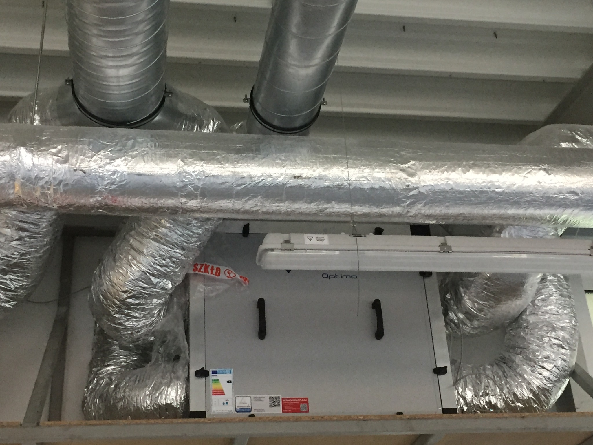 Zamontowana centrala rekuperacyjna Optima z podłączonymi, zaizolowanymi przewodami wentylacyjnymi w obiekcie komercyjnym. Widoczne oznaczenia i etykiety.