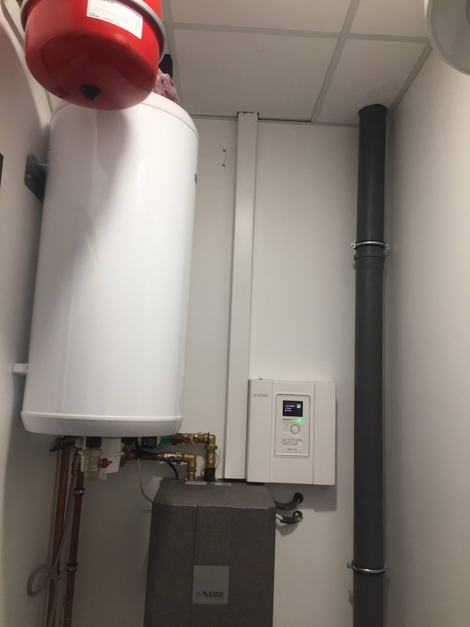 Biały zasobnik ciepłej wody NIBE z czerwonym naczyniem przeponowym, szarą pompą ciepła i panelem sterowania SMO 20 w schludnej kotłowni, rury i instalacje.