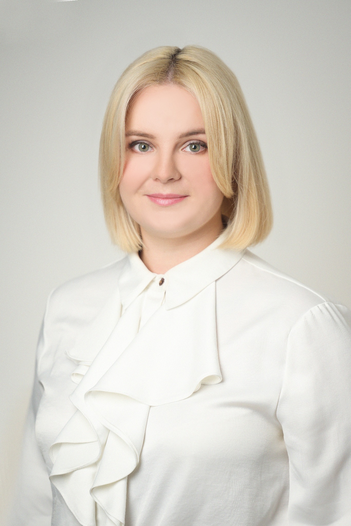 Portret kobiety z blond włosami, ubrana w białą bluzkę z falbanami, na jasnym tle. Delikatny makijaż, profesjonalny wygląd.