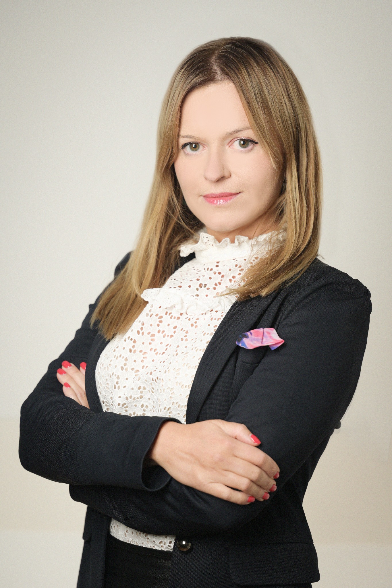 Portret kobiety w czarnej marynarce i koronkowej bluzce, z rękami skrzyżowanymi na piersiach, patrzącej w obiektyw, na neutralnym tle.