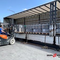 Grupa Dekarska Michał Boguszewski - Pomarańczowy wózek widłowy z operatorem załadowuje palety z betonową dachówką BMI Braas do skrzyni ciężarówki na placu z kostki brukowej.