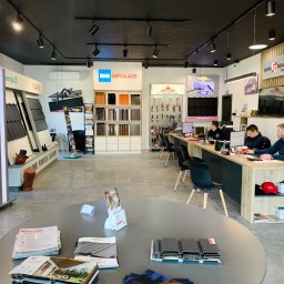 Grupa Dekarska Michał Boguszewski - Wnętrze biura firmy dekarskiej: ekspozycja dachówek różnych marek (BMI Braas, Ruukki, Pruszyński), katalogi na okrągłym stole, pracownicy przy biurku, logo 'Grupa Dekarska Boguszewski' na ścianie.