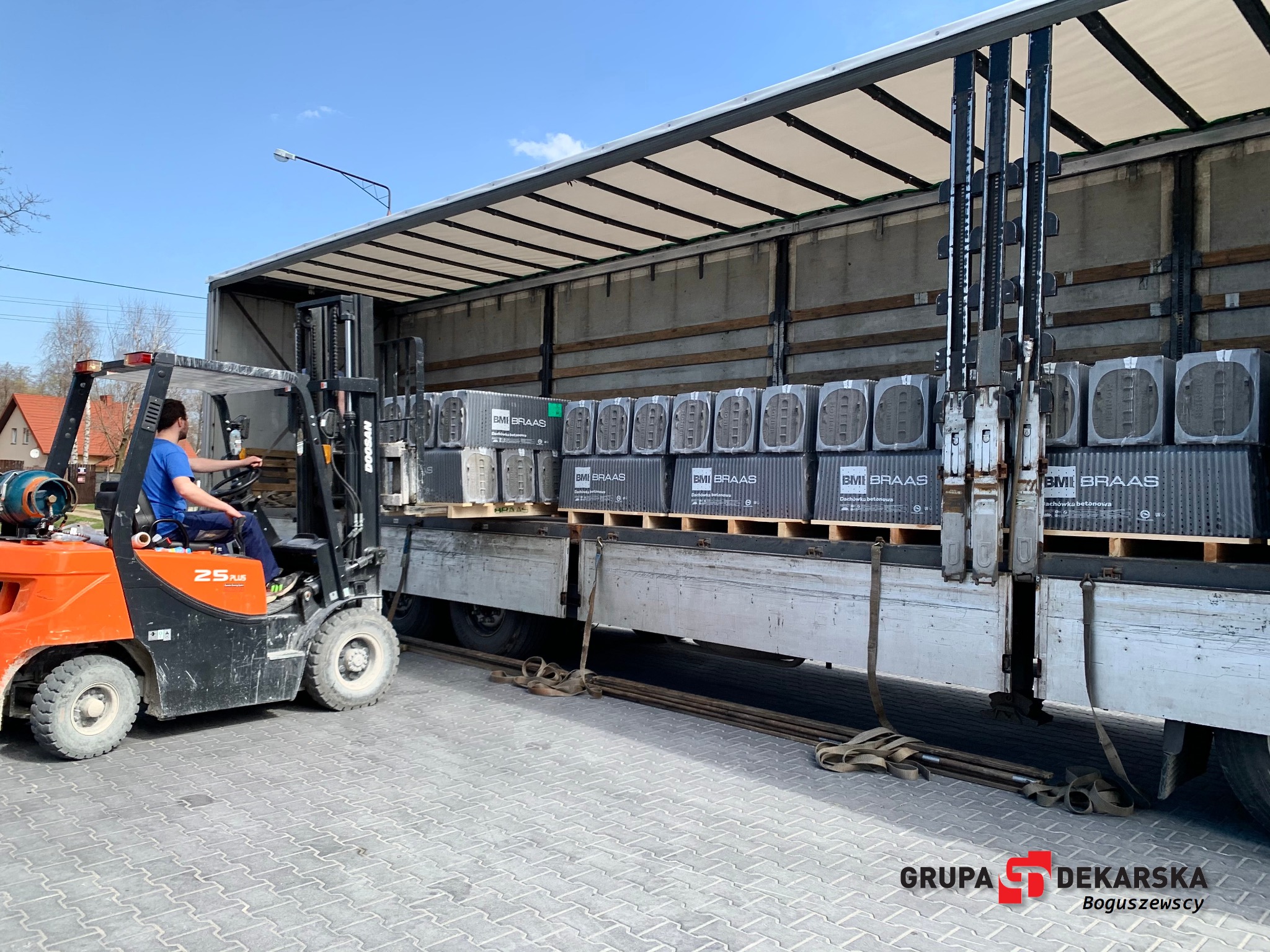 Pomarańczowy wózek widłowy z operatorem załadowuje palety z betonową dachówką BMI Braas do skrzyni ciężarówki na placu z kostki brukowej.