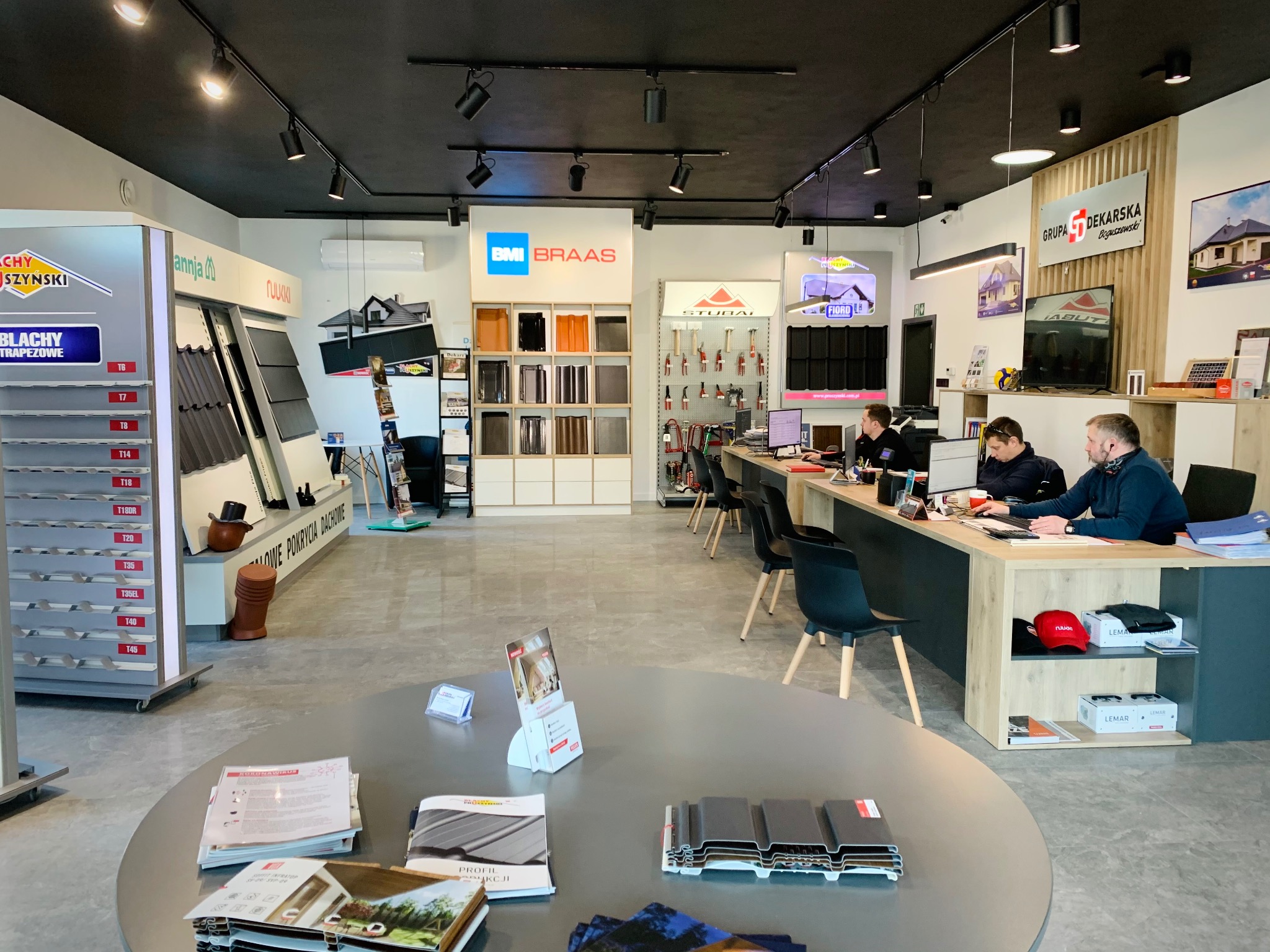 Wnętrze biura firmy dekarskiej: ekspozycja dachówek różnych marek (BMI Braas, Ruukki, Pruszyński), katalogi na okrągłym stole, pracownicy przy biurku, logo 'Grupa Dekarska Boguszewski' na ścianie.