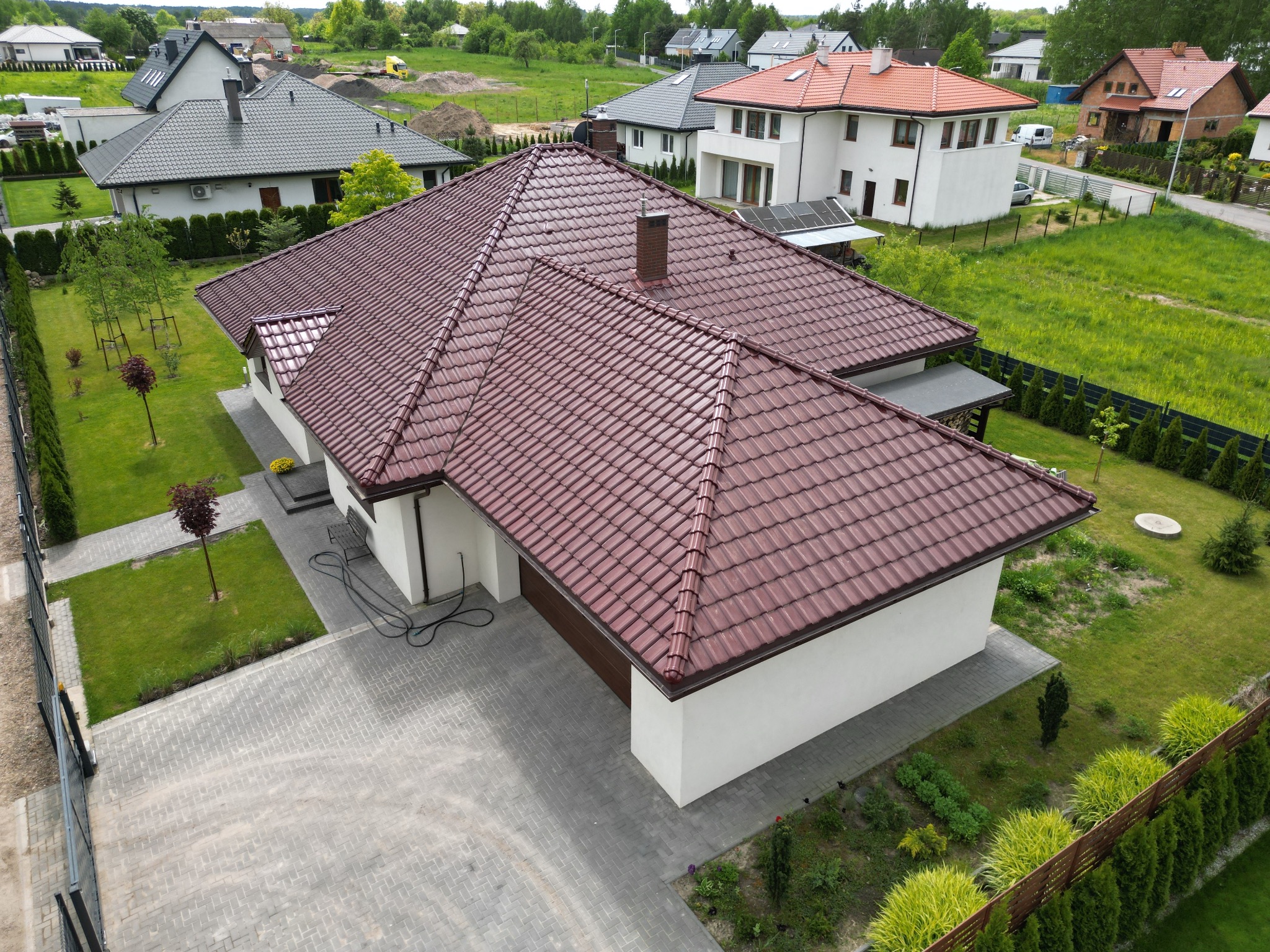 Widok z góry na dom jednorodzinny z nowym, bordowym dachem z dachówki ceramicznej, widoczny komin z cegły i zadbany ogród z kostką brukową.