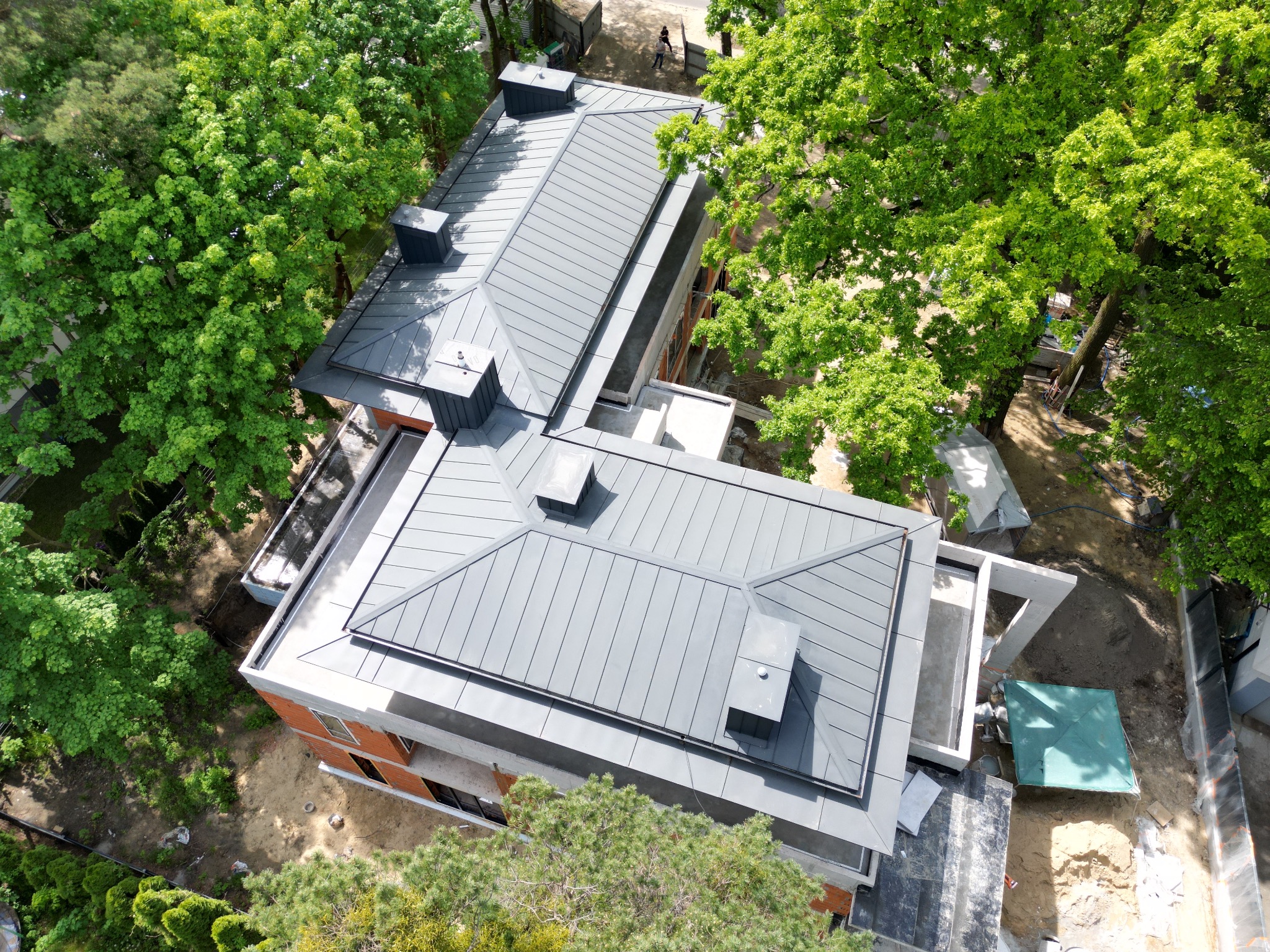 Widok z góry na szary, metalowy dach dwuspadowy z lukarnami na nowym domu w trakcie budowy, otoczonym zielenią drzew.