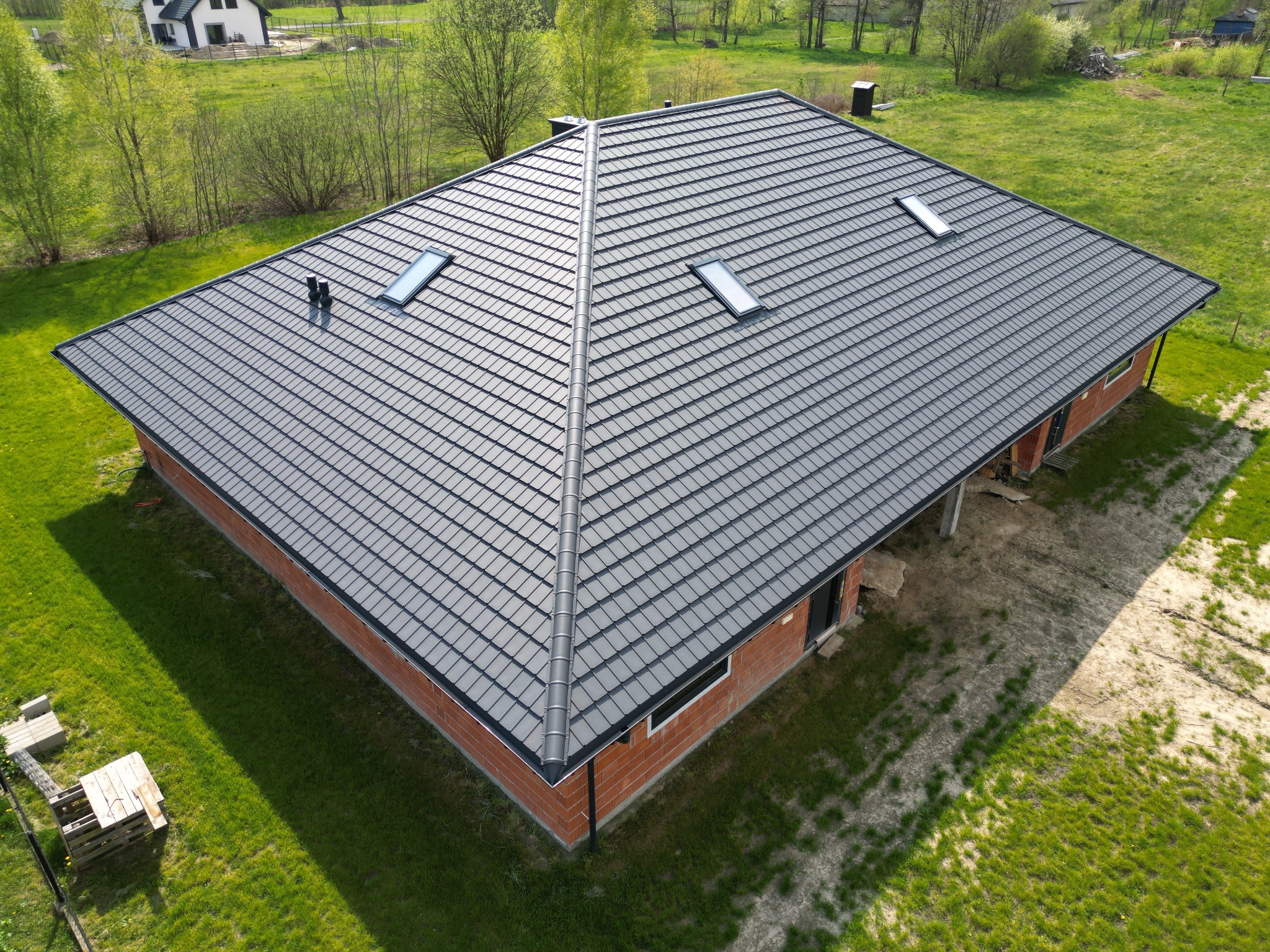 Widok z góry na dom z nowym, ciemnoszarym dachem z dachówki z trzema oknami dachowymi, obróbkami blacharskimi i rynnami, na tle zielonego trawnika i drzew.