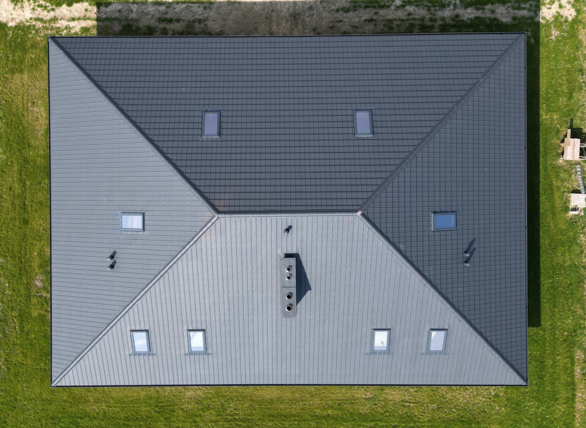 Widok z góry na dwuspadowy dach w Jaktorowie-Kolonii, pokryty częściowo blachą na rąbek stojący, a częściowo dachówką, z oknami dachowymi i kominem.