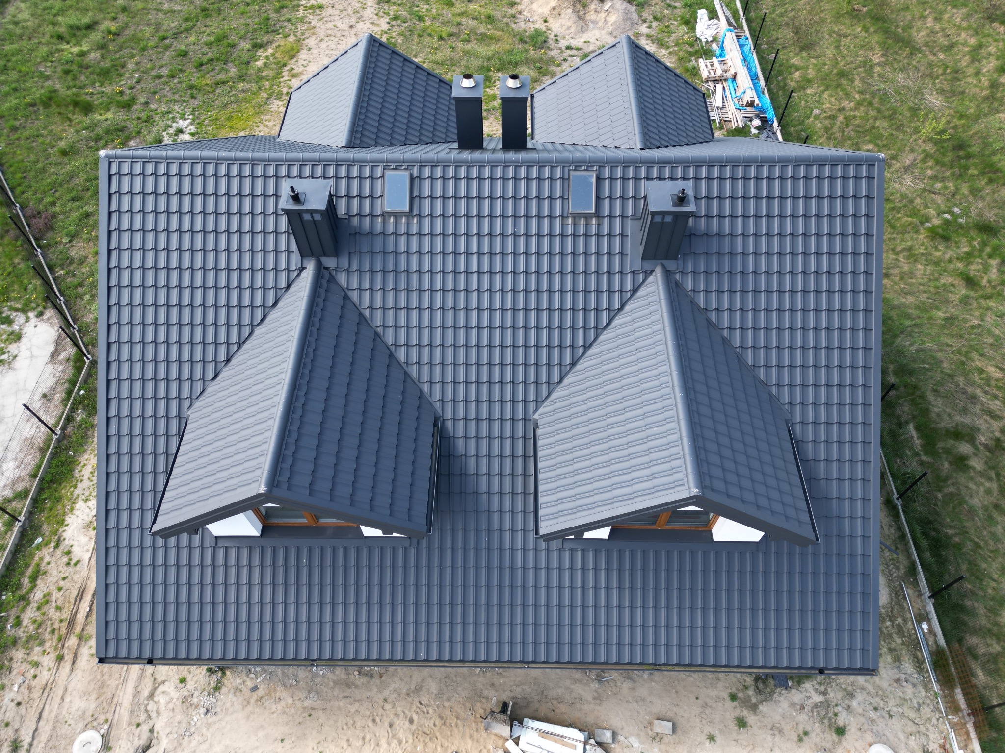 Widok z góry na nowy, grafitowy dach z dachówki modułowej z dwoma kominami i dwoma oknami dachowymi typu lukarna, otoczony zielenią i ogrodzeniem.