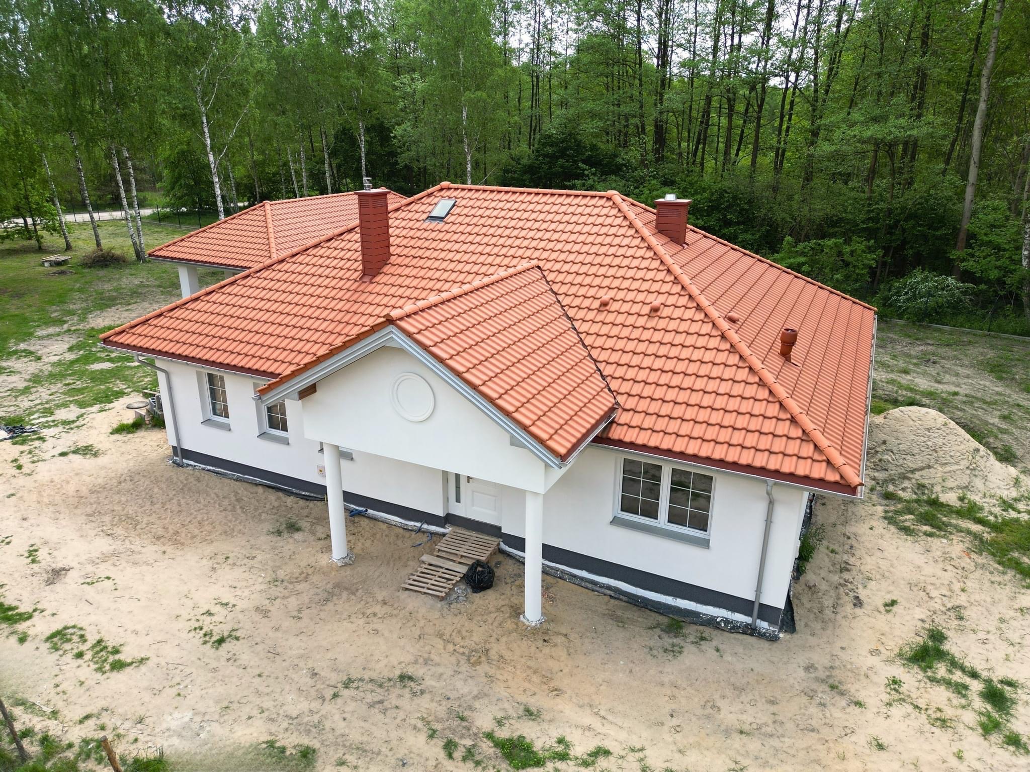 Dom jednorodzinny z nowym, pomarańczowo-czerwonym dachem z dachówki ceramicznej, widok z góry, na tle lasu brzozowego, teren wokół domu częściowo pokryty piaskiem.