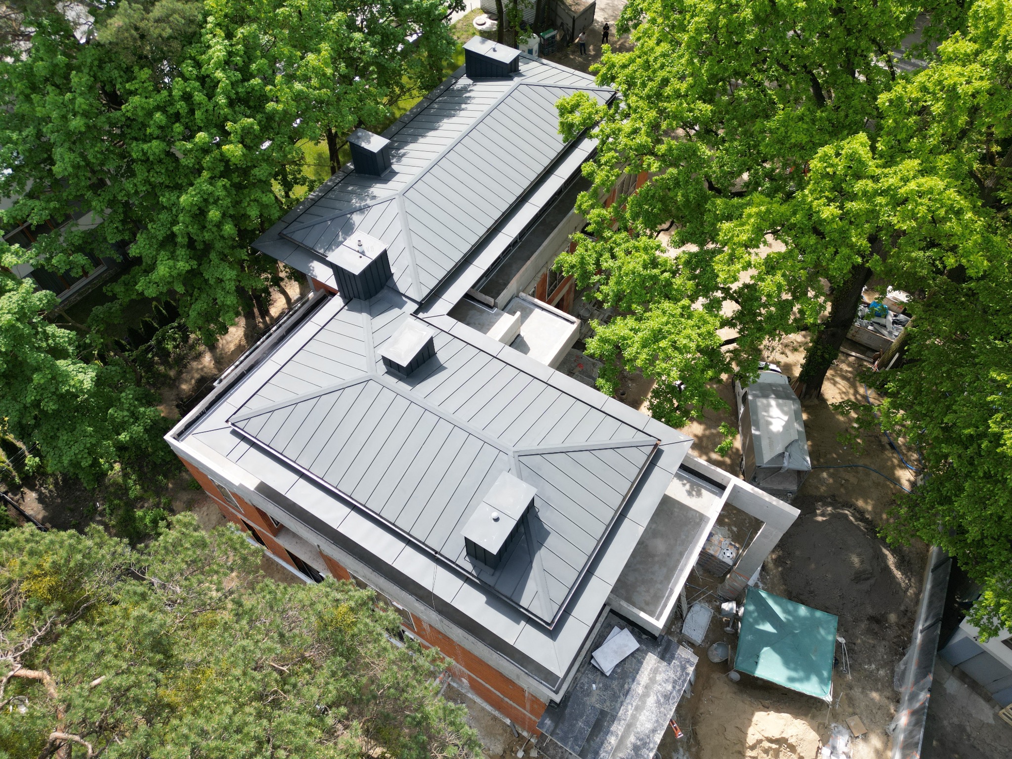 Widok z góry na dwukondygnacyjny budynek z szarym dachem panelowym i kominami, otoczony zielenią drzew, budowa w trakcie wykończenia.