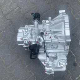 Srebrna skrzynia biegów Yaris II 1.4 D4D leżąca na szarej kostce brukowej, z widocznymi czerwonymi elementami i czarnymi wtyczkami. Na obudowie odręczny napis '06-08'.