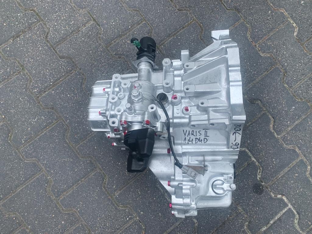 Srebrna skrzynia biegów Yaris II 1.4 D4D leżąca na szarej kostce brukowej, z widocznymi czerwonymi elementami i czarnymi wtyczkami. Na obudowie odręczny napis '06-08'.