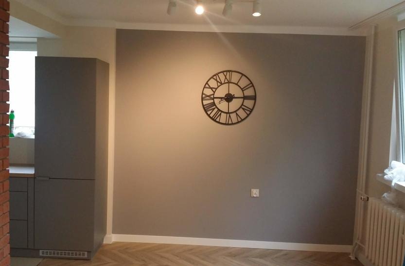 Pomieszczenie po remoncie: szara ściana z dekoracyjnym zegarem, parkiet ułożony w jodełkę, widoczne oświetlenie sufitowe i grzejnik.