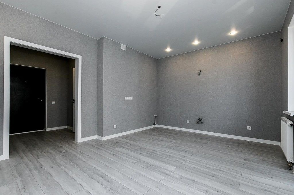 Pusty, szary pokój po remoncie z ułożonymi jasnymi panelami podłogowymi, szpachlowanymi ścianami z widocznymi gniazdkami i punktowym oświetleniem sufitowym. Niedokończone podłączenie lampy sufitowej.