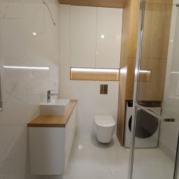 Extra Front s.c - Nowoczesna łazienka z białymi płytkami imitującymi marmur, zabudową meblową z drewnianymi akcentami, umywalką nablatową, toaletą i pralką ukrytą w zabudowie.