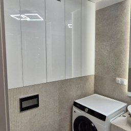 Extra Front s.c - Białe szafki wiszące z trzema poziomymi szczelinami wentylacyjnymi nad pralką w nowoczesnej łazience z beżowymi płytkami i podwieszaną toaletą.