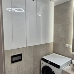 Białe szafki wiszące z trzema poziomymi szczelinami wentylacyjnymi nad pralką w nowoczesnej łazience z beżowymi płytkami i podwieszaną toaletą.