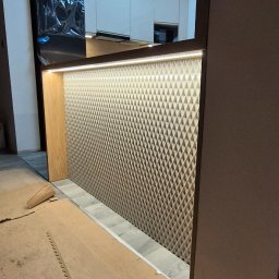 Extra Front s.c - Montaż panelu dekoracyjnego z trójwymiarowym wzorem w kuchni, widoczne zabezpieczenia podłogi i fragment zabudowy kuchennej.