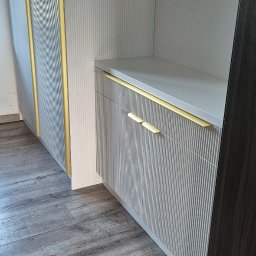 Extra Front s.c - Biała szafa wnękowa z ryflowanymi frontami i złotymi uchwytami, widoczny fragment podłogi z paneli.