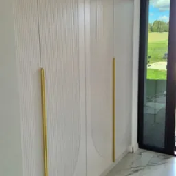 Wysoka szafa z jasnymi frontami, zdobiona pionowymi żłobieniami i motywem półkola, z długimi, złotymi uchwytami, na tle podłogi z marmurowych płytek i widokiem na zewnątrz przez czarne ramy okienne.