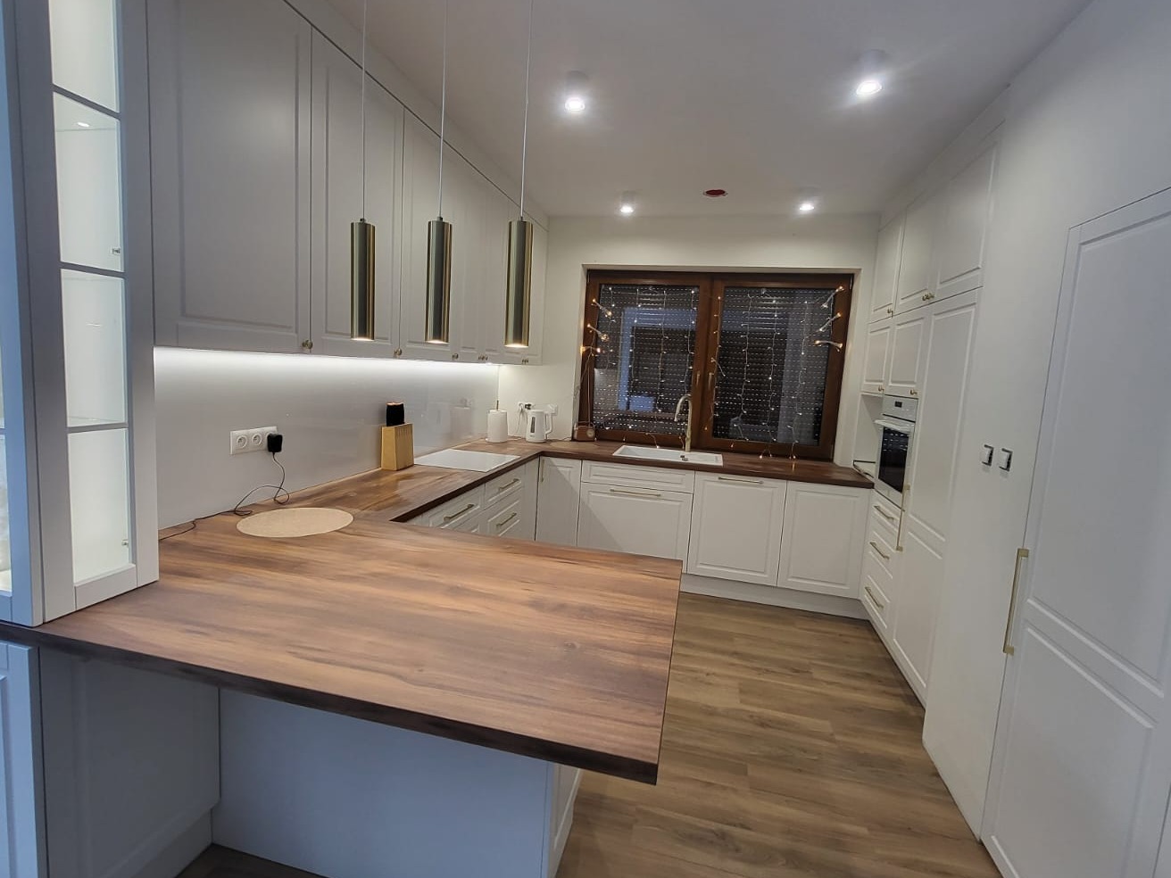 Jasna kuchnia z drewnianym blatem, wyspą i złotymi lampami wiszącymi, oświetlona listwą LED pod szafkami.