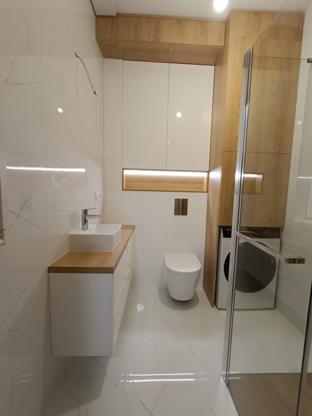 Nowoczesna łazienka z białymi płytkami imitującymi marmur, zabudową meblową z drewnianymi akcentami, umywalką nablatową, toaletą i pralką ukrytą w zabudowie.