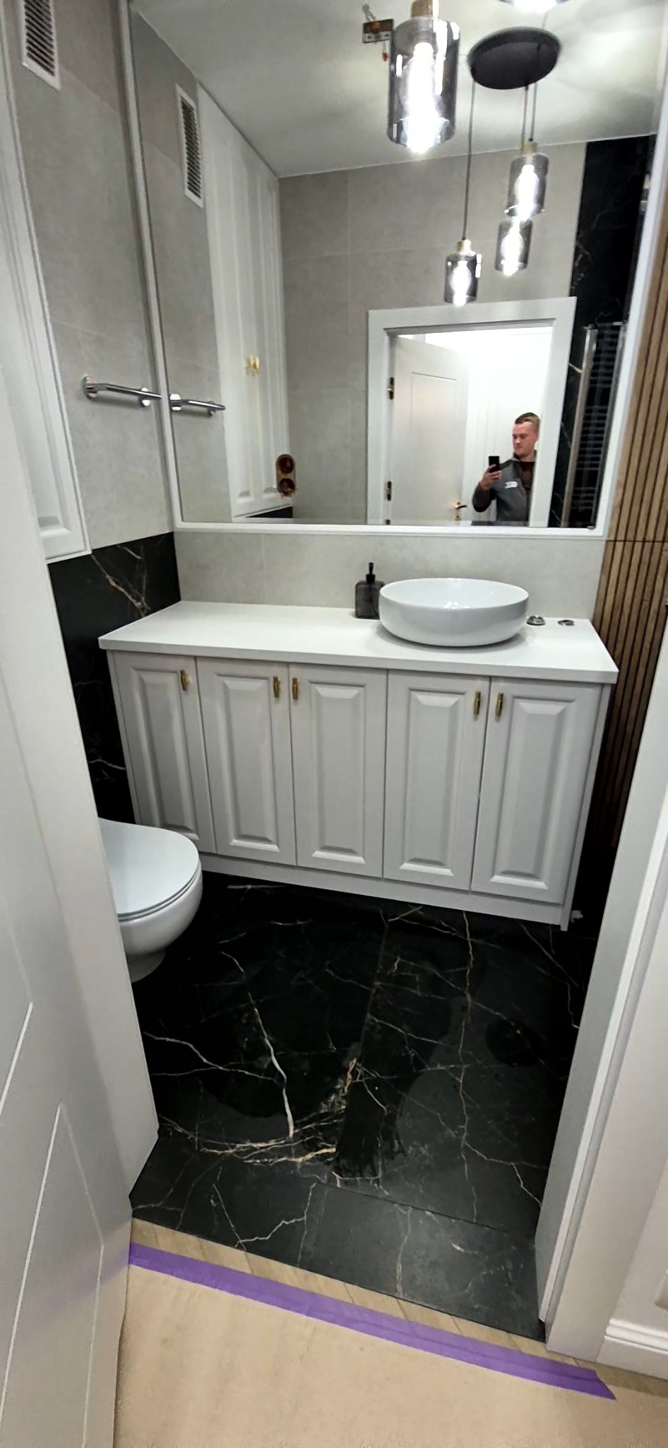 Nowoczesna łazienka z czarną marmurową podłogą, białymi szafkami pod umywalką nablatową, lustrem z odbiciem osoby robiącej zdjęcie i wiszącymi lampami.
