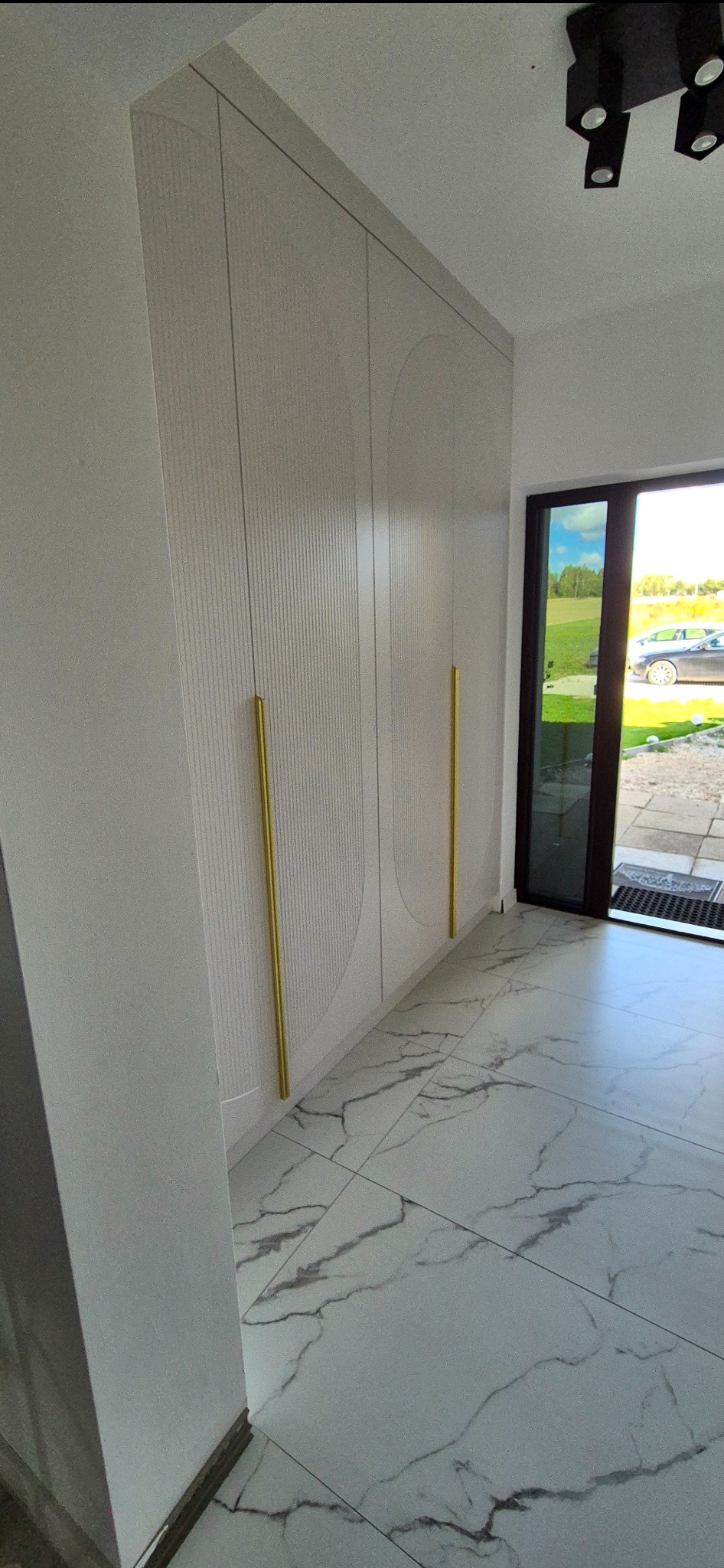 Biała szafa w przedpokoju z frezowanymi drzwiami i złotymi uchwytami, marmurowa podłoga, widok na ogród przez przeszklone drzwi.