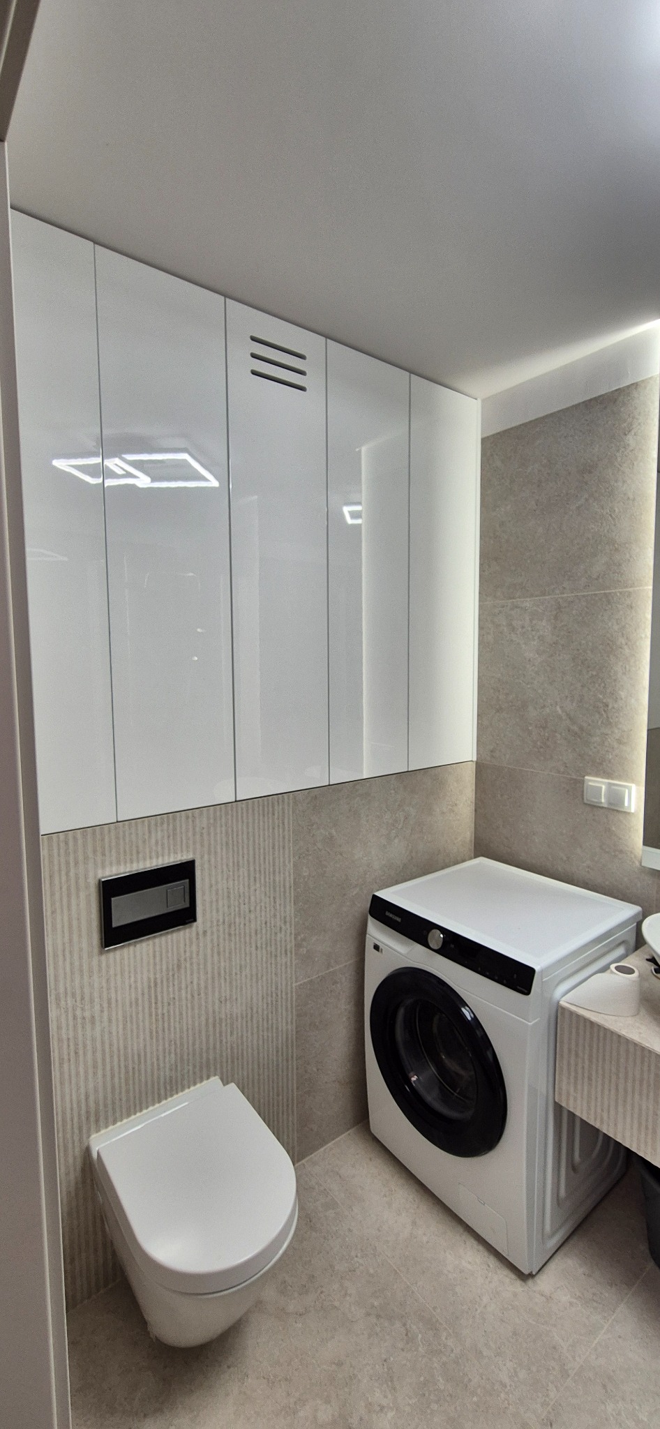 Białe szafki wiszące z trzema poziomymi szczelinami wentylacyjnymi nad pralką w nowoczesnej łazience z beżowymi płytkami i podwieszaną toaletą.