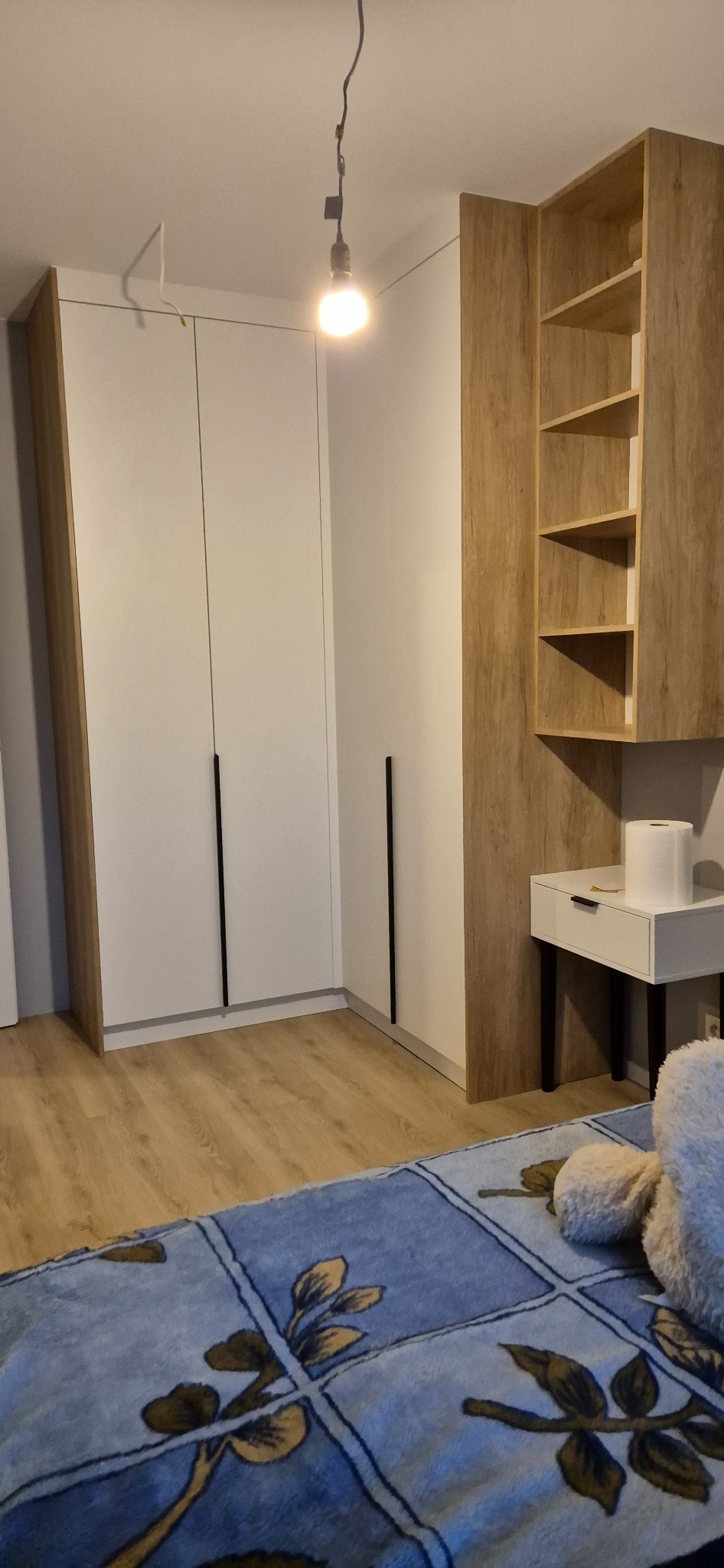 Wnętrze sypialni z zabudową szaf i półkami z drewna, oświetlone pojedynczą żarówką wiszącą na kablu.