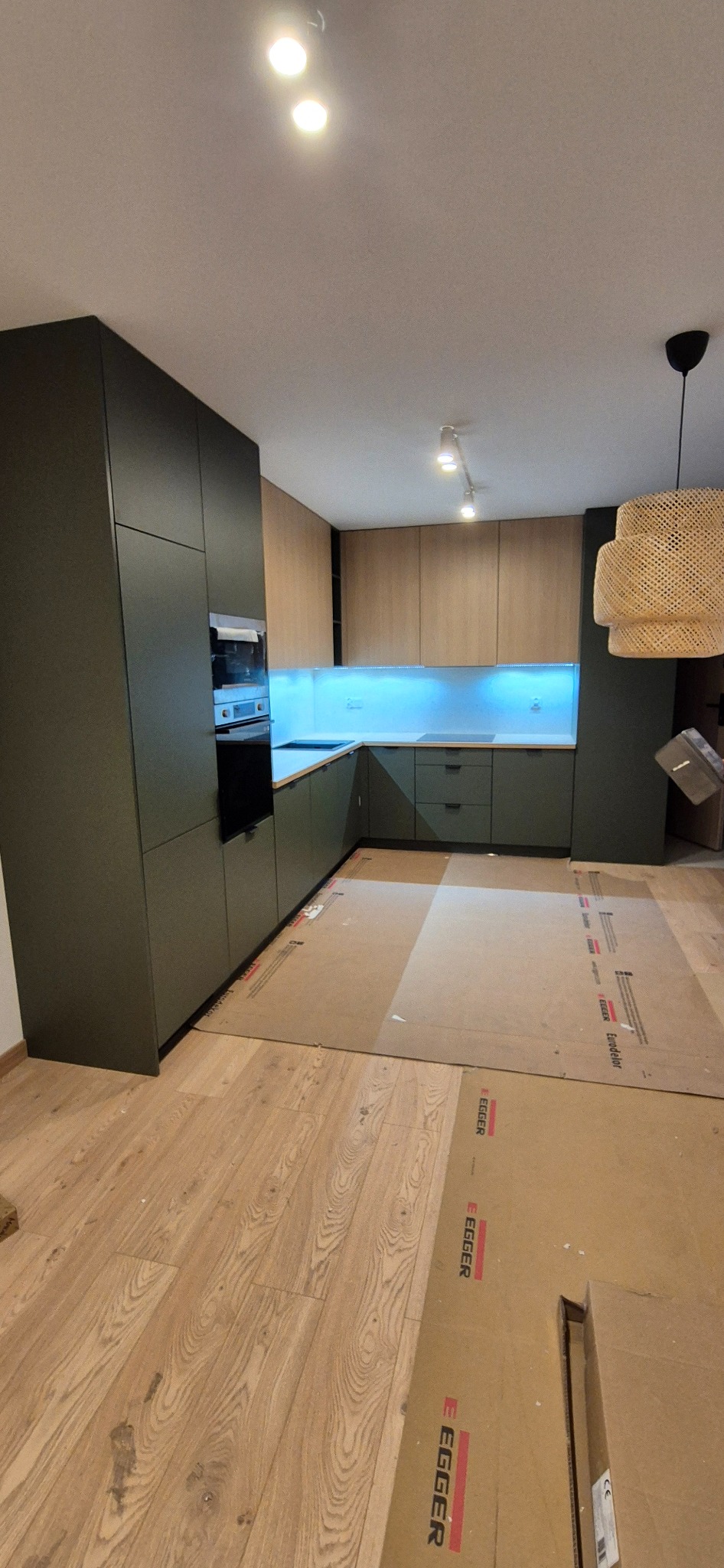 Nowoczesna kuchnia z ciemnozielonymi szafkami i drewnianymi blatami, podświetlona niebieskim światłem, z podłogą zabezpieczoną kartonem podczas montażu.