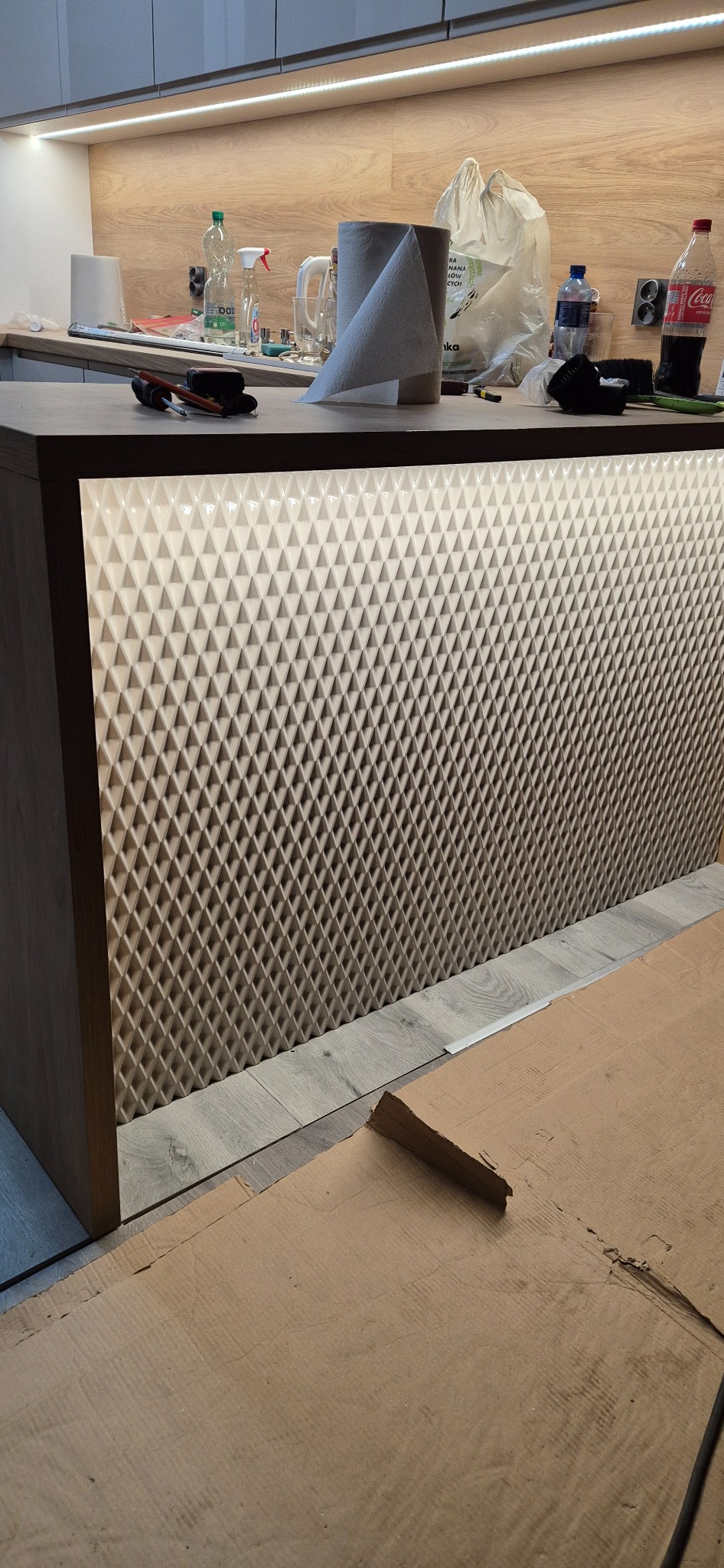 Fragment zabudowy kuchennej z podświetlanym, trójwymiarowym panelem dekoracyjnym, widoczne narzędzia i materiały zabezpieczające podłogę podczas montażu.