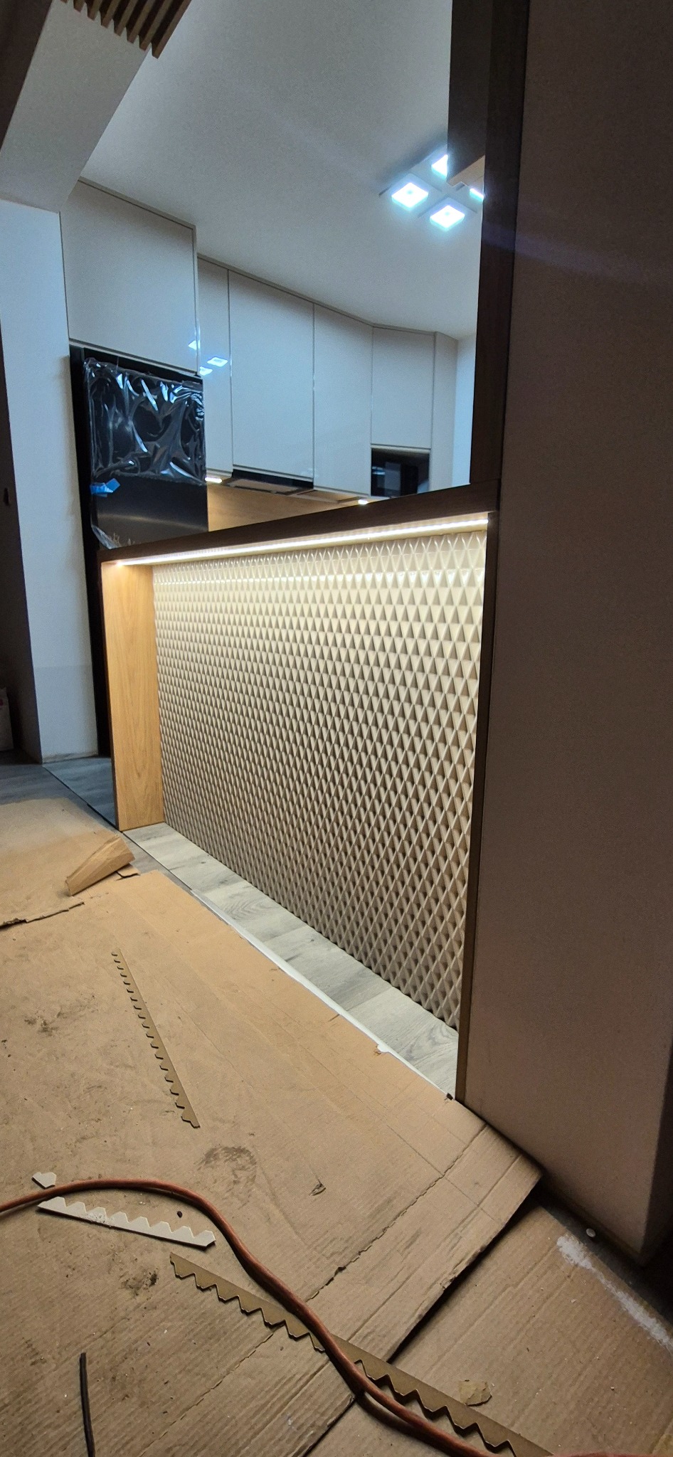 Montaż panelu dekoracyjnego z trójwymiarowym wzorem w kuchni, widoczne zabezpieczenia podłogi i fragment zabudowy kuchennej.