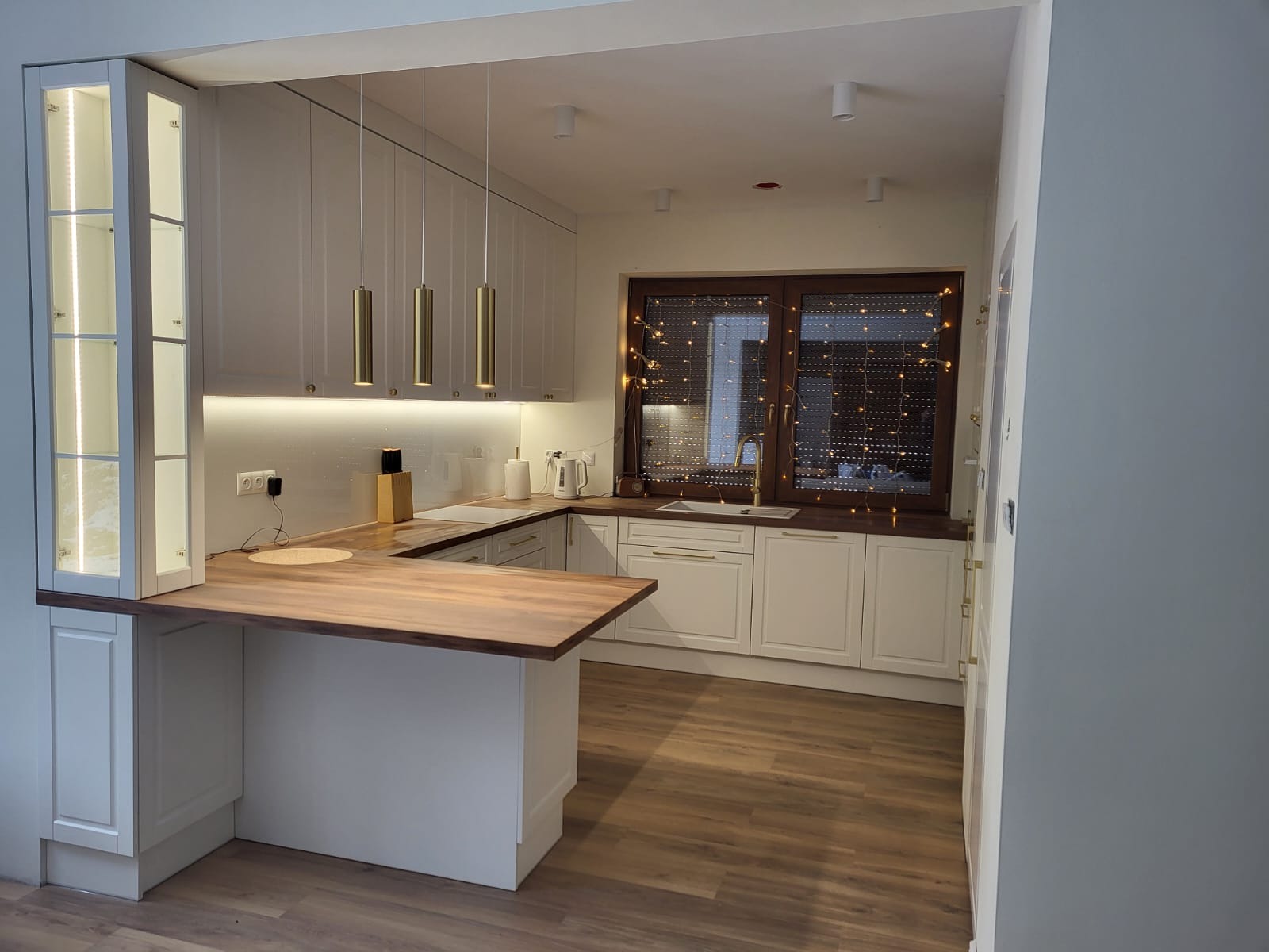 Nowoczesna kuchnia w bieli z drewnianym blatem i złotymi lampami wiszącymi, okno z dekoracyjnymi światełkami.