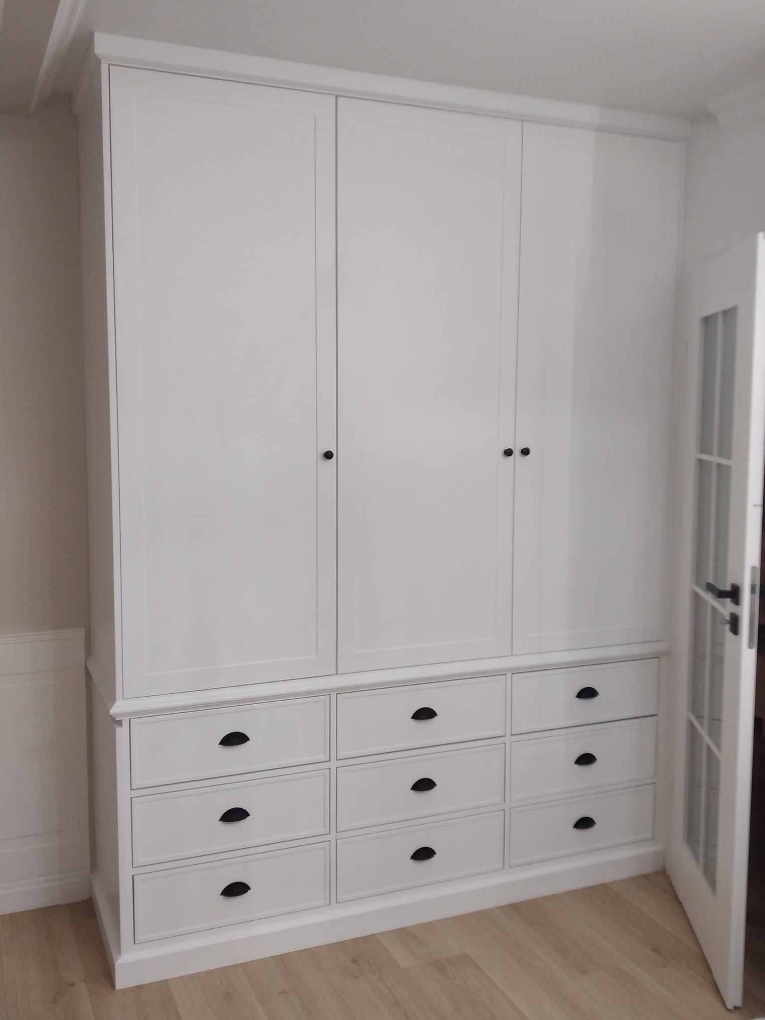 Biała szafa z dziewięcioma szufladami i trzema drzwiami, czarne okrągłe uchwyty, widoczna ościeżnica białych drzwi z szybami