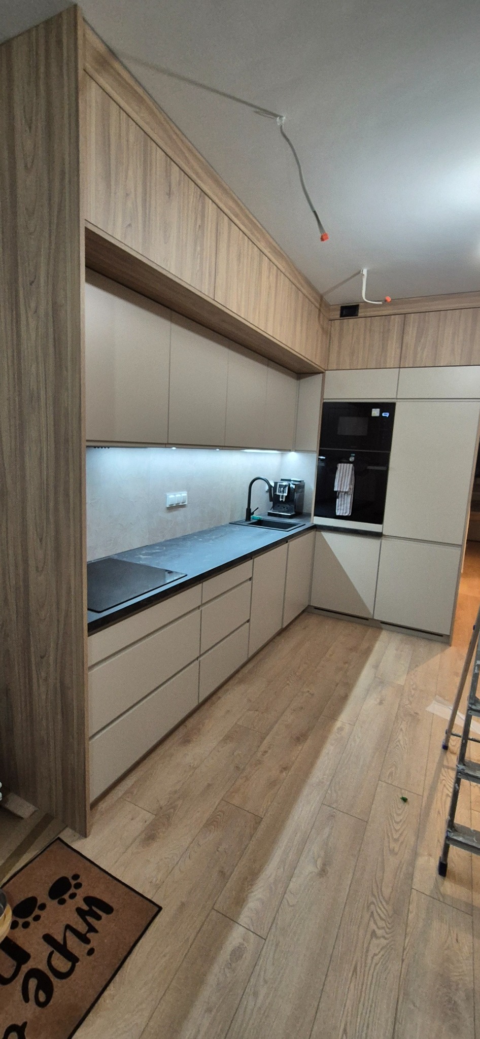 Nowoczesna kuchnia z beżowymi szafkami i blatem w kolorze antracytu, oświetlenie LED pod szafkami, drewniana podłoga, widoczne niedokończone podłączenie elektryczne na suficie i drabina malarska.