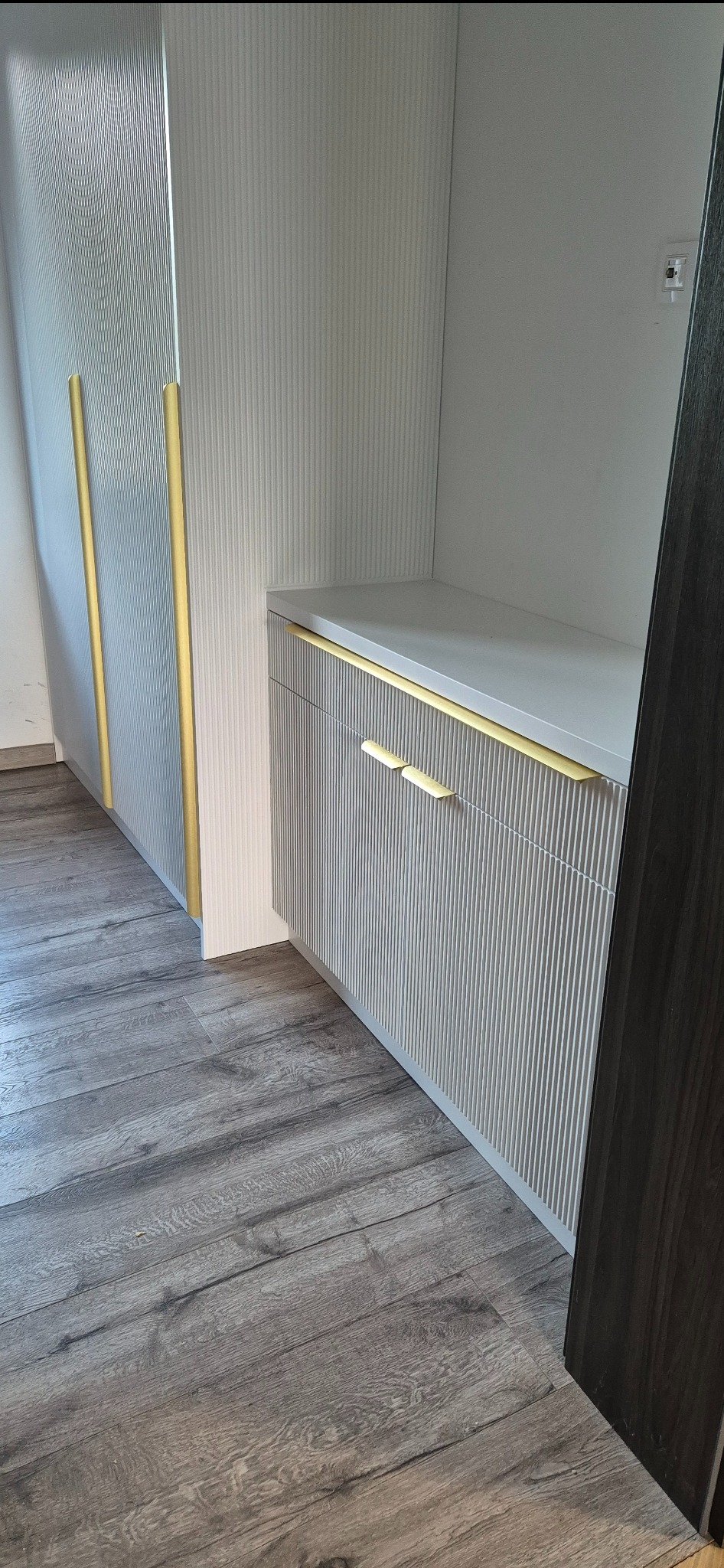 Biała szafa wnękowa z ryflowanymi frontami i złotymi uchwytami, widoczny fragment podłogi z paneli.