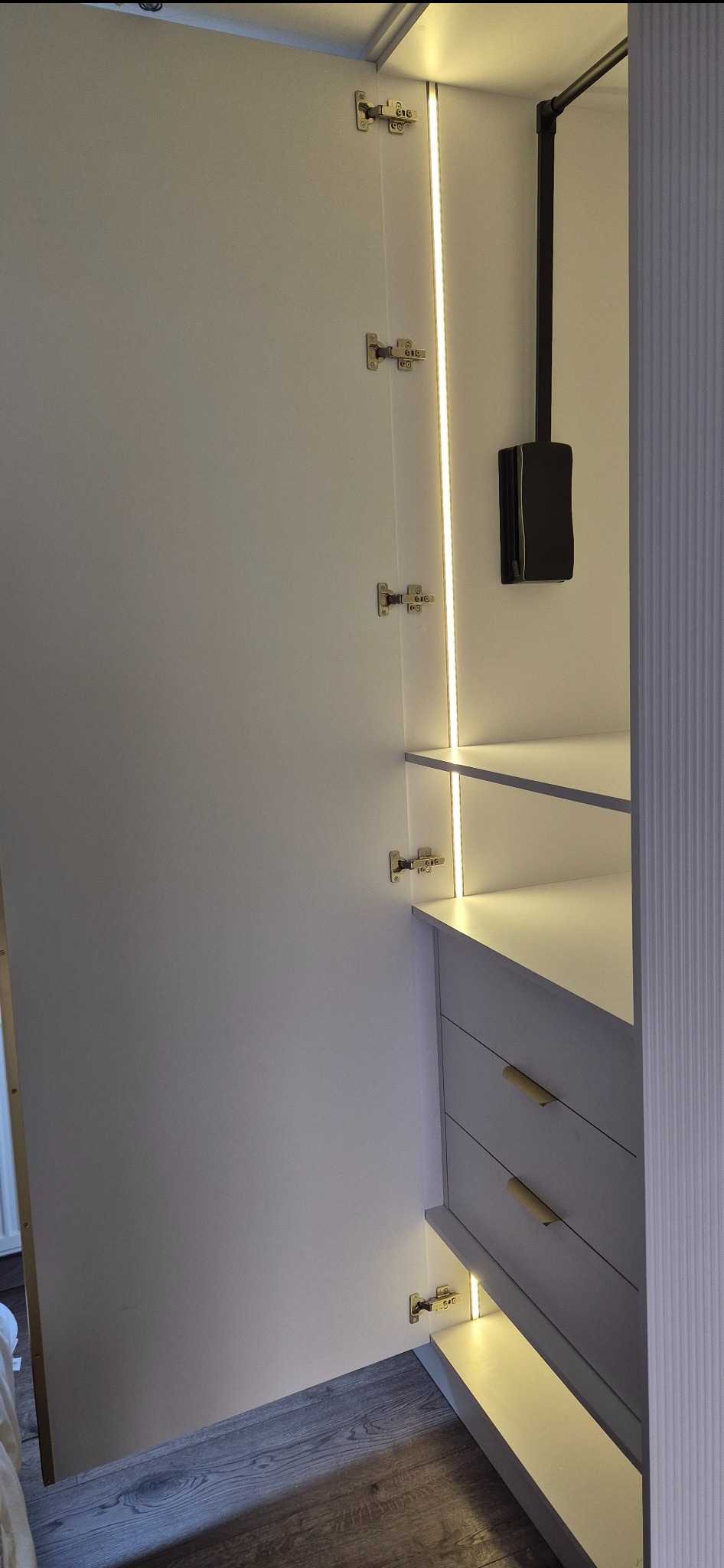 Otwarta szafa wnękowa z szufladami, oświetleniem LED i drążkiem na ubrania, widoczne zawiasy i drewniana podłoga.