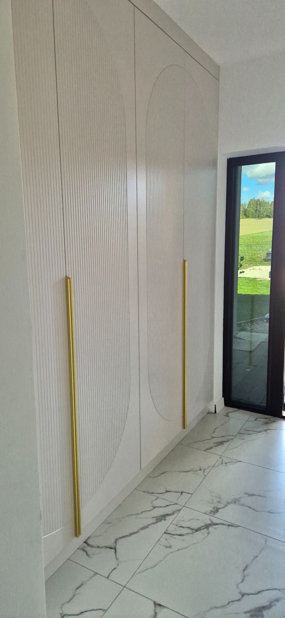 Wysoka szafa z jasnymi frontami, zdobiona pionowymi żłobieniami i motywem półkola, z długimi, złotymi uchwytami, na tle podłogi z marmurowych płytek i widokiem na zewnątrz przez czarne ramy okienne.