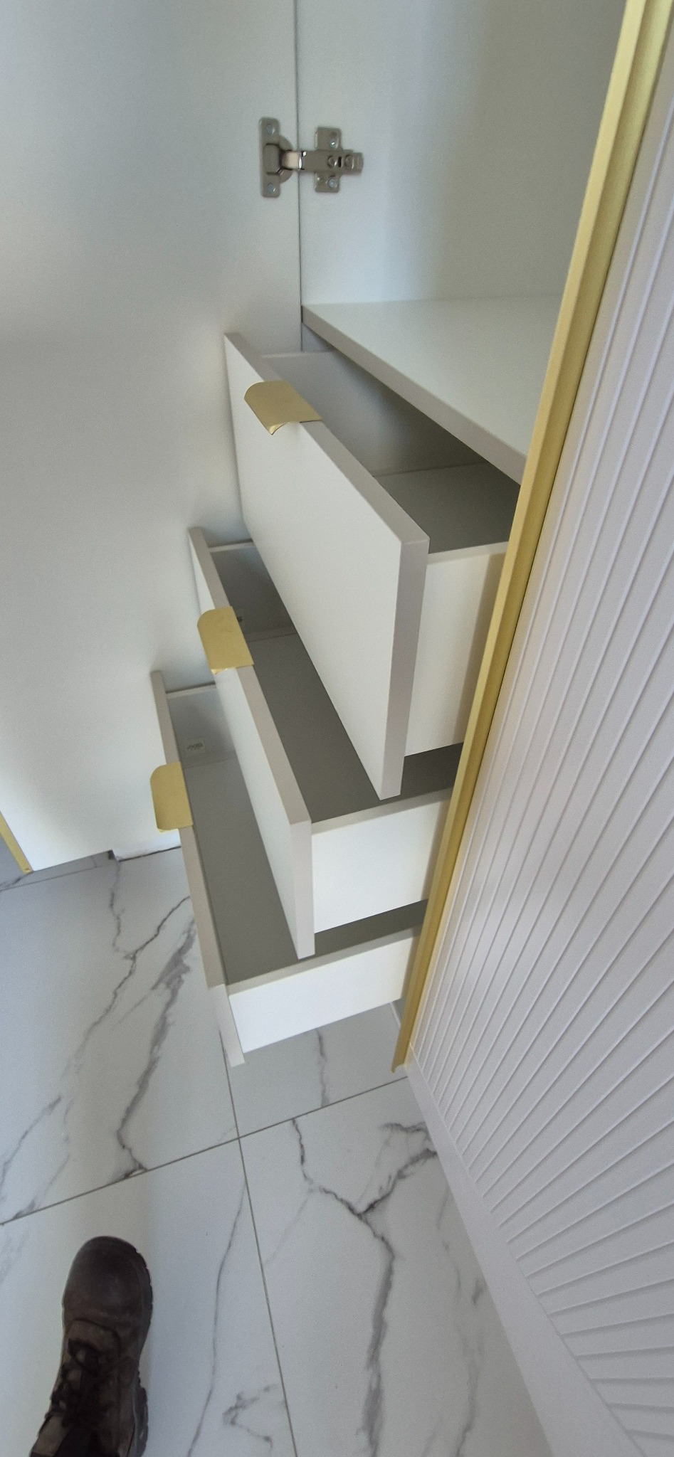 Wnętrze szafy z trzema wysuwanymi szufladami z prostymi, złotymi uchwytami, widok z góry na marmurowe płytki podłogowe i fragment buta.